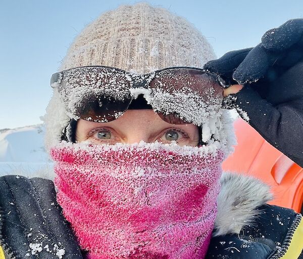 A lady wears a pink face buff, beanie and sunglasses perched on her head, all covered in a thick layer of ice crystals.