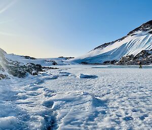 Sea-ice with a foamy looking covering of ice. There are snow-covered hills and bright blue sky in the background.