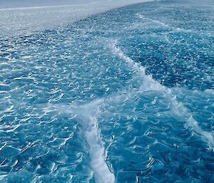 Rippled blue lake ice that looks like small waves.  A snow covered hill is visible in the background.