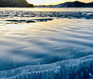 Vertically rising air bubbles seen through frozen blue lake ice. Hills surrounding the lake visible in the background.