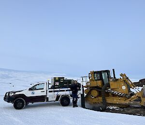 A white utility is backed up to a yellow dozer. The landscape is covered in snow and ice and there is a rocky slope visible in the right of frame.