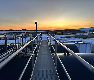 A metal walkway above large fuel tanks is in the centre of frame. There is an orange sunrise on the horizon with rocky islands and frozen sea visible.