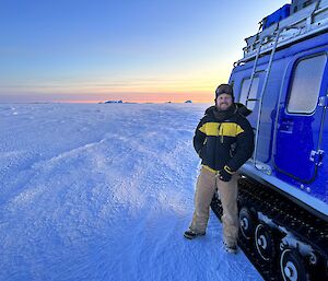 A man is standing next to a blue Hägglunds vehicle on a ice covered sea. He is smiling broadly at the camera.