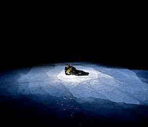 A man is laying relaxed on a frozen lake at night. There is a glowing light coming from under him inside the ice.