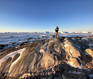 A man is standing on top of a rocky hilly island. In the distance are a large number of icebergs locked into the frozen sea.
