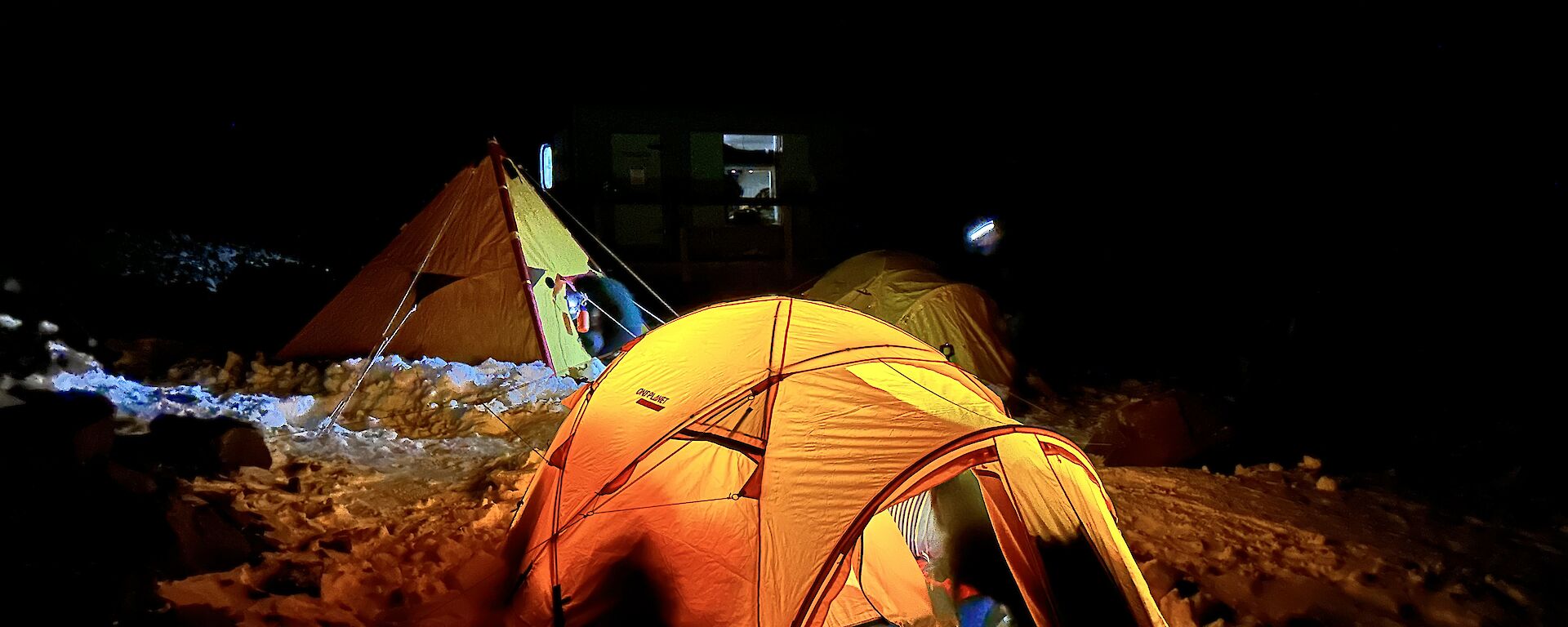 A yellow brightly lit tent is setup on the snow with a yellow pyramid tent and another dome tent in the background.