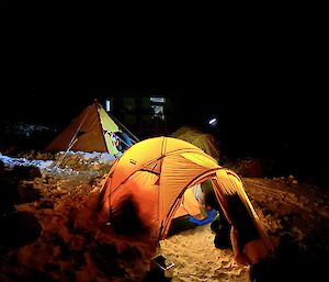 A yellow brightly lit tent is setup on the snow with a yellow pyramid tent and another dome tent in the background.