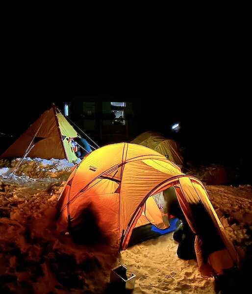 A yellow brightly lit tent is setup on the snow with a yellow pyramid tent and another dome tent in the background.
