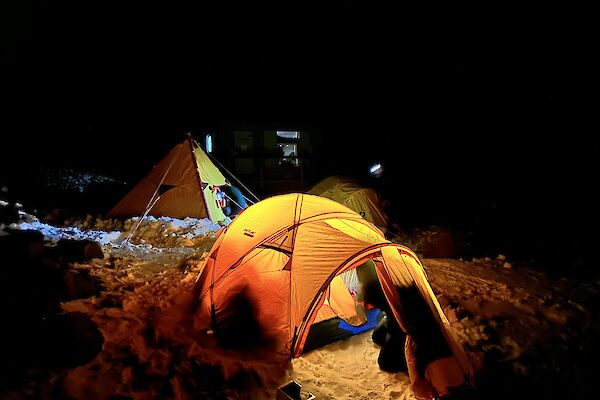 A yellow brightly lit tent is setup on the snow with a yellow pyramid tent and another dome tent in the background.