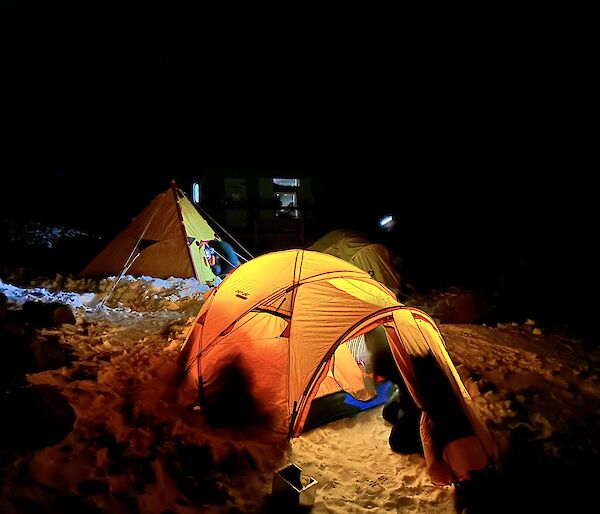 A yellow brightly lit tent is setup on the snow with a yellow pyramid tent and another dome tent in the background.