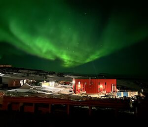 A bright green aurora is in the night sky above a number of buildings that are brightly lit.