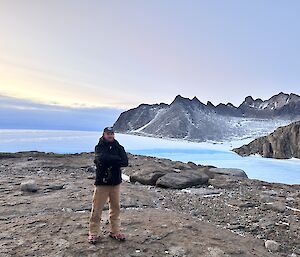 A man is standing on a rocky landscape smiling at the camera. In the background is an ice covered plateau and a rocky mountain ridge rising in the distance.
