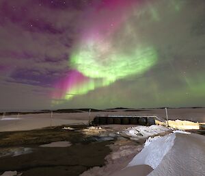 A bright green and purple aurora is lighting up clouds in the night sky over a frozen harbour. In the foreground are a number of large fuel tanks and a snow covered, rocky, landscape.