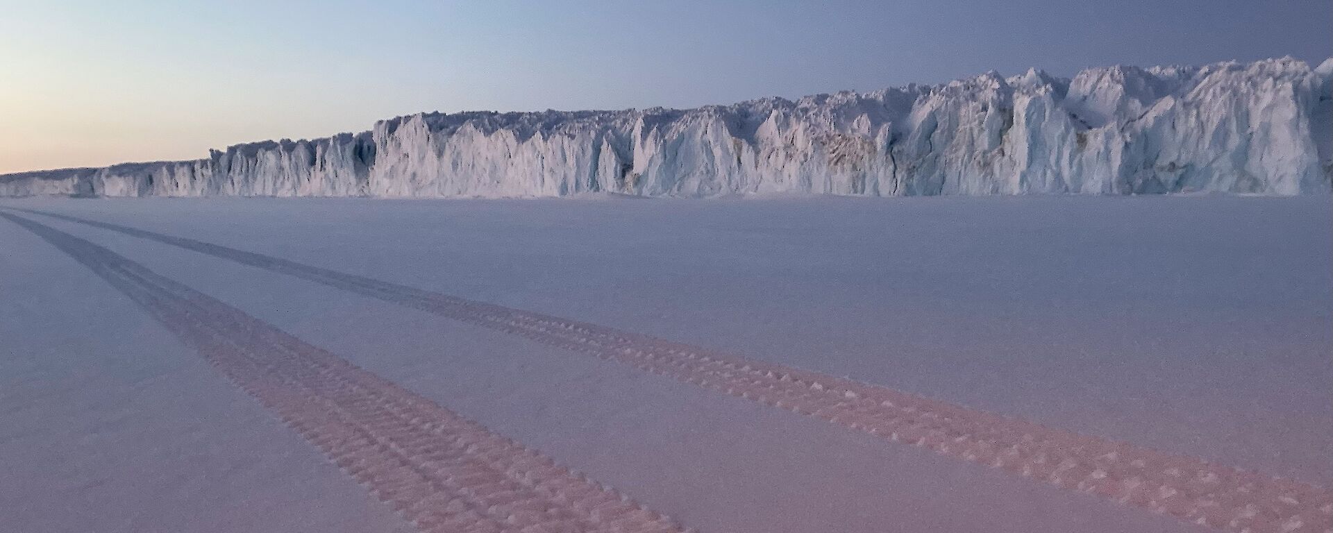 Vehicle tracks in fresh snow with the side wall of a glacier visible in the background.