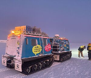Two people drilling into the sea-ice in front of a blue Hägglunds vehicle.