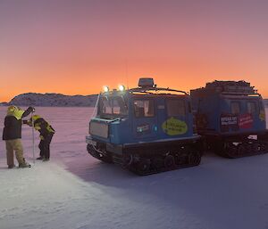 Two people drilling into the sea-ice in front of a blue Hägglunds vehicle. The sky is a pale pinky red.