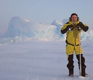 A man wearing yellow protective clothing standing on the sea-ice with a long drill. In the distance behind him is a large white iceberg.