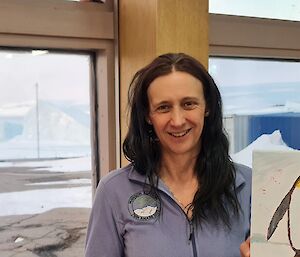 A woman is holding a canvas painting of an emperor penguin. Through the windows behind her there are two large icebergs visible beyond a rocky middle ground.