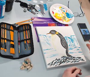 A canvas painting of an emperor penguin and icebergs is on a table surrounded by paints and paintbrushes.