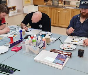 Three men are painting on canvas at a table. There are a lot of paints and brushes on the table.