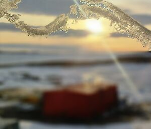 A line of ice is visible across a pane of glass. Beyond the glass a blurred image of a red building and a rocky, snow and ice covered landscape is visible. The sun is setting in the distance.