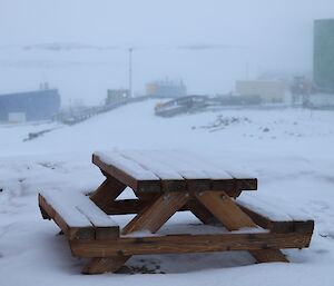 A picnic table and the landscape around it is coated in a layer of fluffy snow. In the background are a number of colourful buildings.