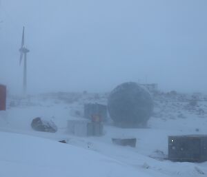 A rocky landscape is covered in a layer of snow. Snow is still falling. There is a wind turbine in the background and a large satellite dome in the centre of frame.