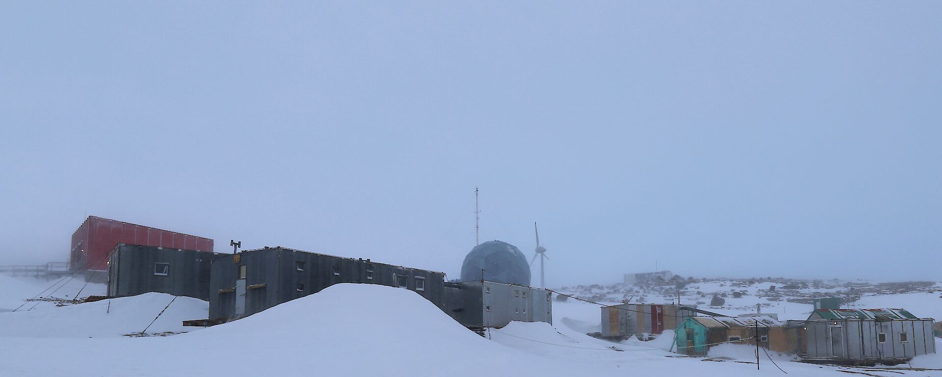 A number of buildings are on a rising rocky landscape that is covered in snow. There are snow drifts against a number of the buildings. There is a wind turbine and large satellite dome in the background.