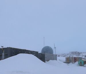 A number of buildings are on a rising rocky landscape that is covered in snow. There are snow drifts against a number of the buildings. There is a wind turbine and large satellite dome in the background.
