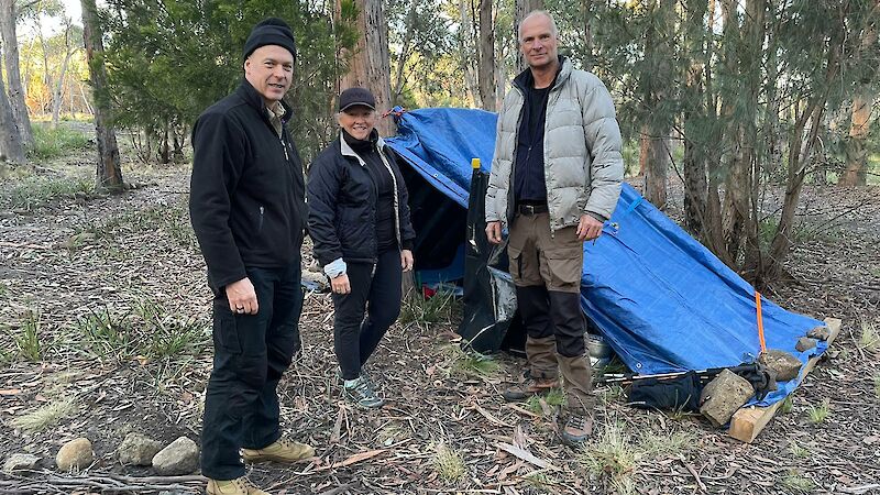 Two men with faces blurred and a woman stand in front of a tarpaulin