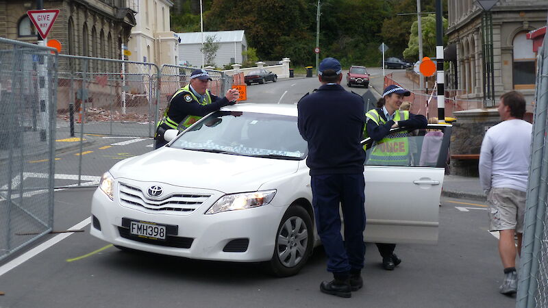 Two policemen and one policewoman stand by a white car in a suburban street talking to a man in a white t-shirt and shorts