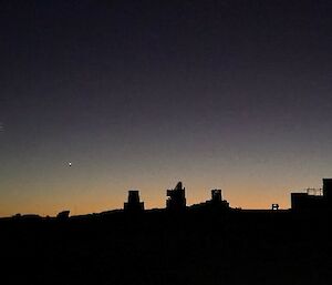 A silhouette of station machinery against a pre-dawn sky. The moon is visible to the left of the machinery.