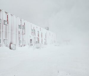 There is a two story metal building that is almost completely covered in snow. The ground is covered in snow and the surrounding air is filled with blowing snow.