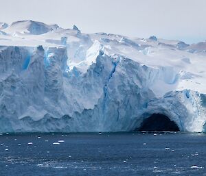 Tall white-blue ice cliffs at the edge of an ice shelf with a dark cave-like hollow at the base where the ocean has melted the ice