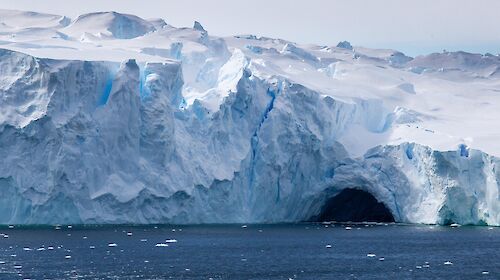 Tall white-blue ice cliffs at the edge of an ice shelf with a dark cave-like hollow at the base where the ocean has melted the ice