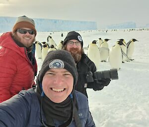 Three men are smiling at the camera. Just behind them are a number of large emperor penguins and in the distance are large iceberg frozen into the sea ice and many more penguins.