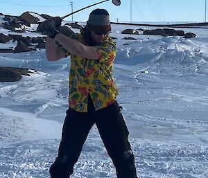 A man in a brightly coloured shirt is about to hit a yellow golf ball. He is standing on snow covered ice and there are tall aerials in the background