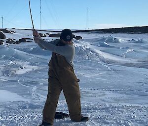 A man in tan overalls and beanie is about to hit a pink golf ball with a golf club. He is standing on an ice covered landscape.