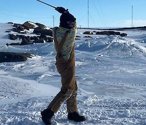 A man in overalls and a brightly coloured shirt has taken a swing at a red golf ball and missed. He is standing on an ice covered landscape.