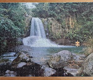 A large jigsaw puzzle of a waterfall is complete on a table however there are two pieces missing.