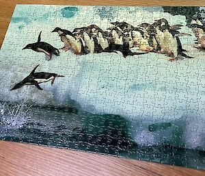 A large jigsaw puzzle of penguins diving into water from an iceberg is complete on a table.