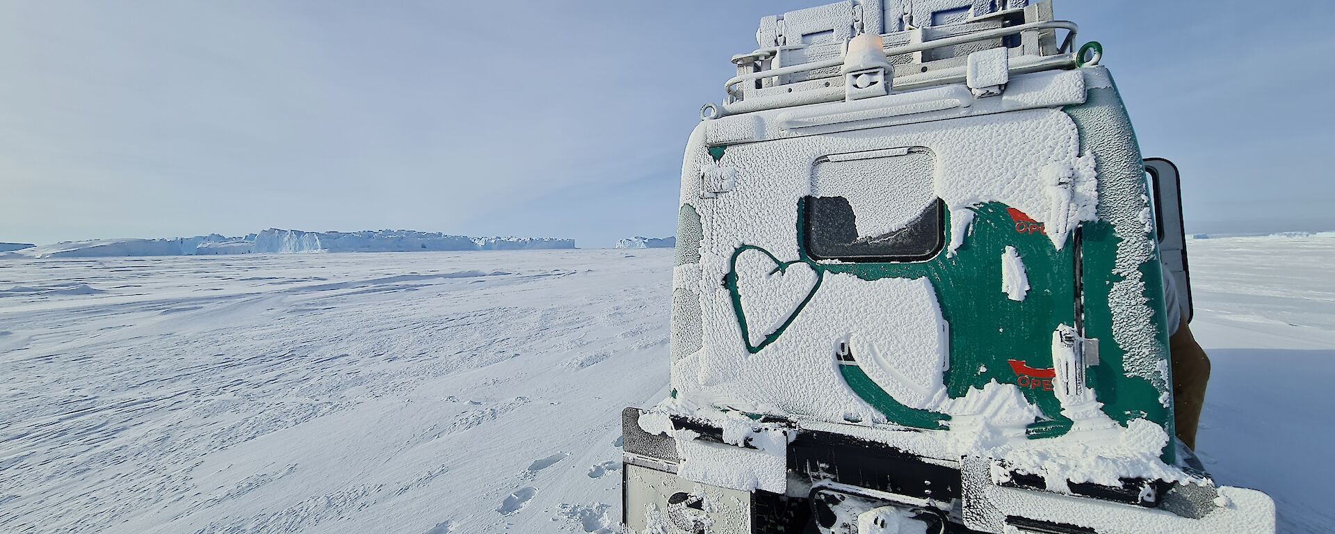 A green Hägglunds vehicle is covered in snow and a heart has been drawn in the snow on the back. It is parked on an expanse of sea ice with large icebergs in the distance