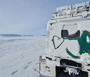 A green Hägglunds vehicle is covered in snow and a heart has been drawn in the snow on the back. It is parked on an expanse of sea ice with large icebergs in the distance