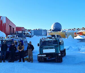 A group of six men are standing in front of two Hägglunds vehicles smiling at the camera. In the background are a number of brightly coloured buildings, a satellite dome and a wind turbine.