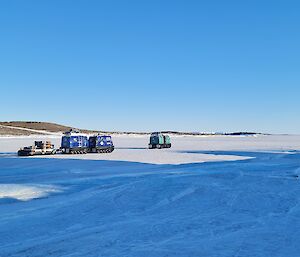 Two Hägglunds vehicles with one pulling a sled are driving away over an ice covered harbour.