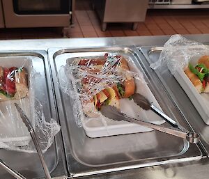 Three trays of sandwiches are on a bain-marie. Tongs are on each tray for service.