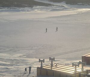 A person is standing next to a golf cart on the edge an ice covered harbour. Two people are on the ice close to a golf flag.