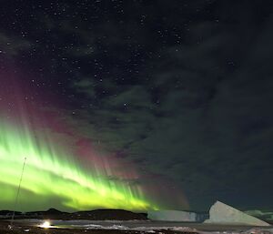 A brightly coloured green and purple aurora is visible in the night sky above two large icebergs and a rocky island.
