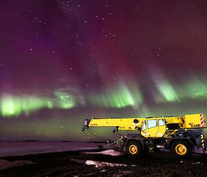 A brightly coloured green and purple aurora is visible in the night sky a bright yellow crane. In the background is an ice covered harbour.