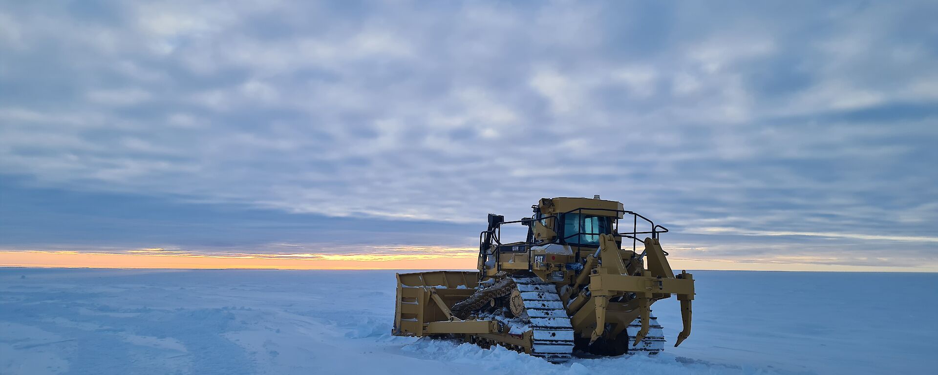 A large yellow bulldozer parked on a flat expanse of snow covered ice. There is an even layer of low cloud stretching out to the horizon.